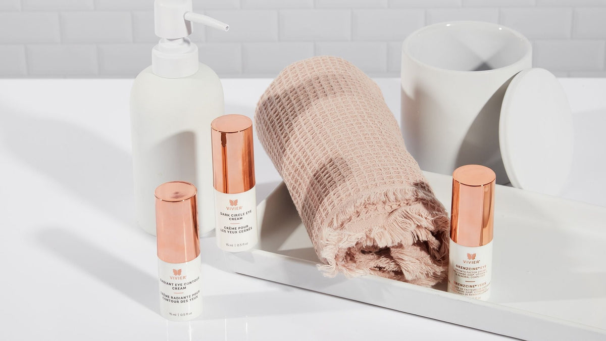Three Vivier eye creams displayed on a bathroom countertop beside a folded towel and skincare accessories.