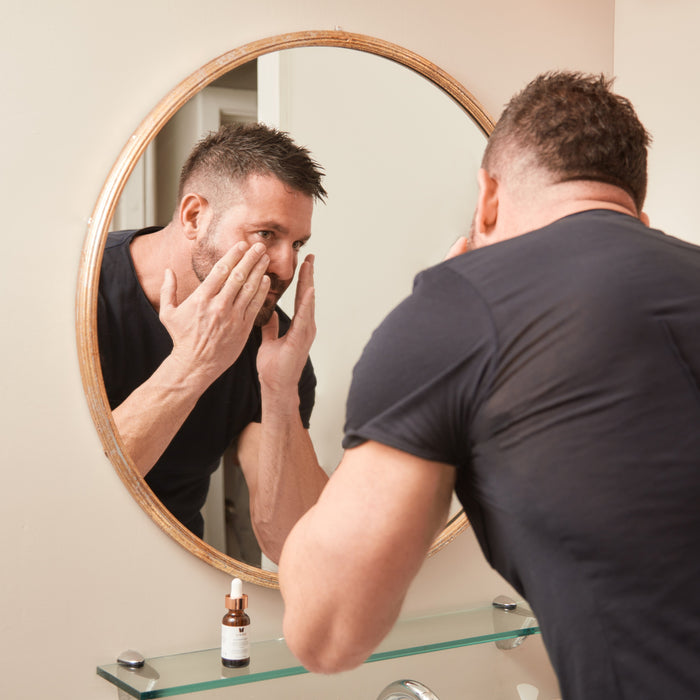Man applying skincare around his eyes while looking into a round bathroom mirror. A Vivier serum bottle sits on a glass shelf below the mirror, highlighting a daily skincare routine in a clean, modern bathroom setting.