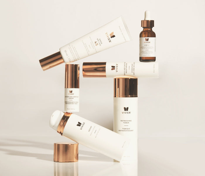 Alt text: Vivier skincare products arranged in a balanced display against a clean white background, featuring sunscreen, vitamin C serum, toner, eye cream, and cleansing products with signature rose gold packaging.